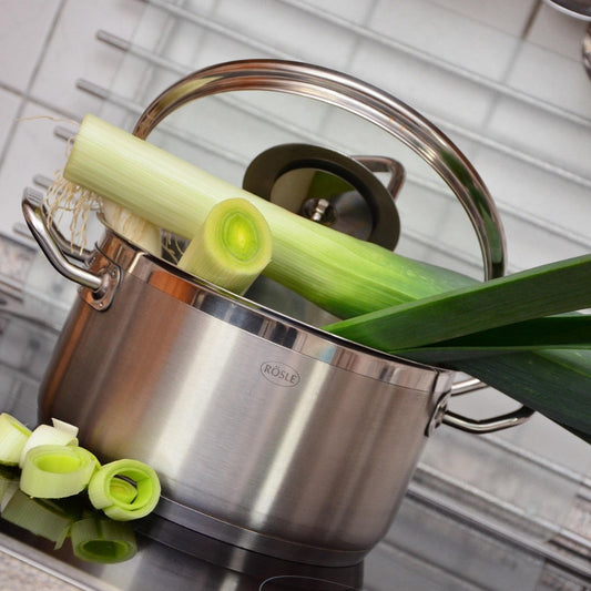 A pot on a stovetop filled with water and containing leeks, with some leeks also placed around the pot, indicating they have been recently washed or are being prepared for cooking.